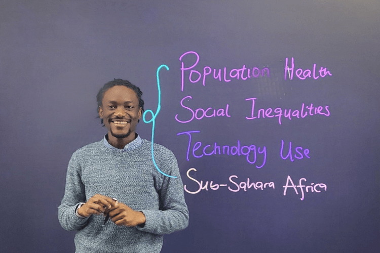 Dr Emmanuel Olamijuwon stands smiling behind a see-through board, on which he has written 'Population Health', 'Social Inequalities', 'Technology Use', 'Sub-Sahara Africa' in purple and pink ink